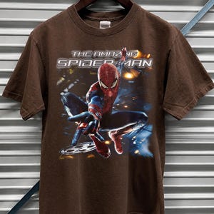 May include: Brown t-shirt featuring the words "THE AMAZING SPIDER-MAN" in white and silver lettering. The graphic shows Spider-Man in a dynamic pose, with a blue and red costume. The shirt is hanging on a wooden hanger.