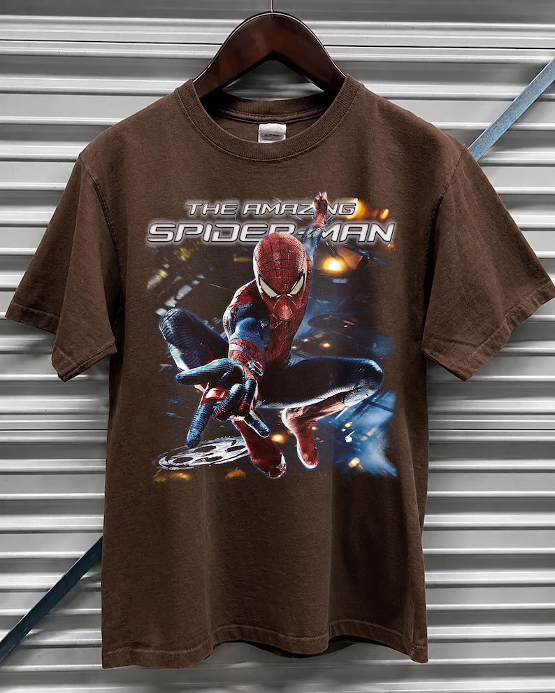 May include: Brown t-shirt featuring the words "THE AMAZING SPIDER-MAN" in white and silver lettering. The graphic shows Spider-Man in a dynamic pose, with a blue and red costume. The shirt is hanging on a wooden hanger.