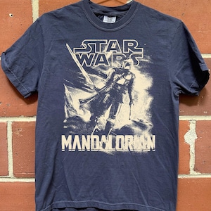 Pu&ograve; includere: T-shirt blu navy con una grafica del Mandaloriano di Star Wars. Il logo di Star Wars &egrave; sopra l'immagine e la parola "MANDALORIAN" &egrave; sotto. La maglietta &egrave; appesa a una gruccia di metallo contro un muro di mattoni.
