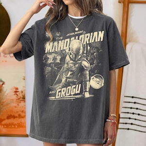 Pu&ograve; includere: T-shirt grigio scuro con grafica vintage di The Mandalorian e Grogu di Star Wars. Il design include il testo "The Mandalorian" e "Grogu" in stile vintage. La maglietta ha maniche corte e una vestibilit&agrave; ampia.