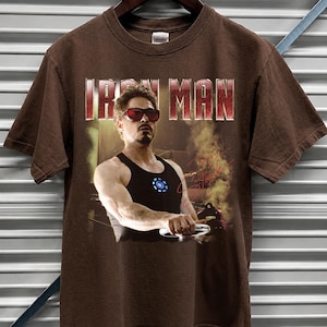 May include: Brown t-shirt featuring the text "IRON MAN" in a red and white font. The shirt displays a graphic of Tony Stark in a black tank top and red sunglasses. The shirt is hanging on a wooden hanger.