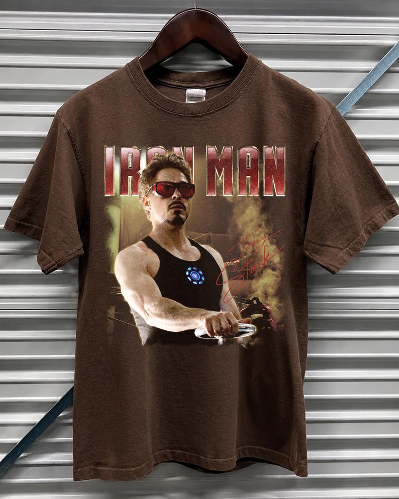 May include: Brown t-shirt featuring the text "IRON MAN" in a red and white font. The shirt displays a graphic of Tony Stark in a black tank top and red sunglasses. The shirt is hanging on a wooden hanger.