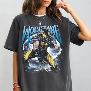 Könnte beinhalten: Dunkelgraues T-Shirt mit einem Wolverine-Motiv. Das Design zeigt das Wort "Wolverine" in Blau und Weiß, mit einer Illustration der Figur in Gelb und Blau. Das T-Shirt hat kurze Ärmel und einen Rundhalsausschnitt.