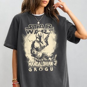 Puede incluir: Camiseta gris oscuro con un gr&aacute;fico de Star Wars. El dise&ntilde;o incluye el texto "STAR WARS" sobre una ilustraci&oacute;n de The Mandalorian, con "THE MANDALORIAN AND GROGU" debajo. La imagen tiene un aspecto vintage.