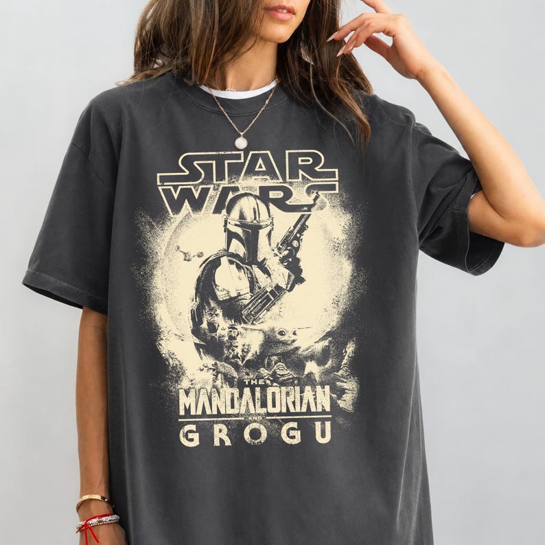 Puede incluir: Camiseta gris oscuro con un gr&aacute;fico de Star Wars. El dise&ntilde;o incluye el texto "STAR WARS" sobre una ilustraci&oacute;n de The Mandalorian, con "THE MANDALORIAN AND GROGU" debajo. La imagen tiene un aspecto vintage.