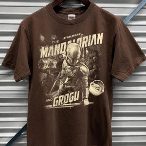 Pu&ograve; includere: T-shirt marrone a maniche corte con una grafica di The Mandalorian e Grogu di Star Wars. Il design include il testo "The Mandalorian" e "Grogu" in uno stile vintage. La maglietta &egrave; appesa a una gruccia di legno.