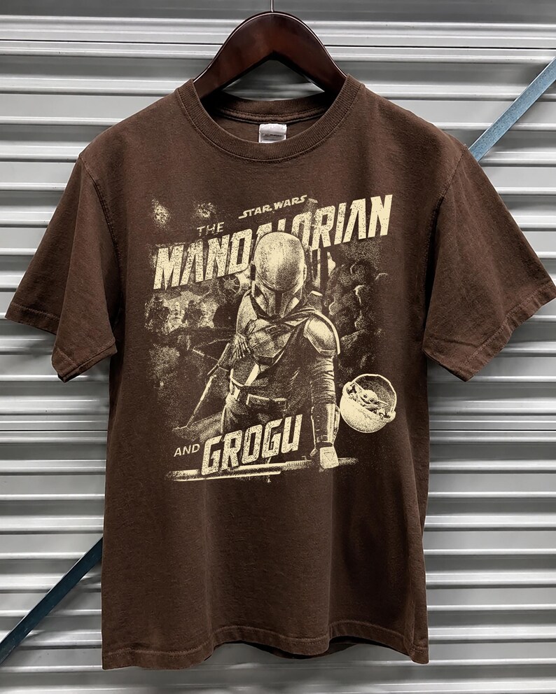 Pu&ograve; includere: T-shirt marrone a maniche corte con una grafica di The Mandalorian e Grogu di Star Wars. Il design include il testo "The Mandalorian" e "Grogu" in uno stile vintage. La maglietta &egrave; appesa a una gruccia di legno.