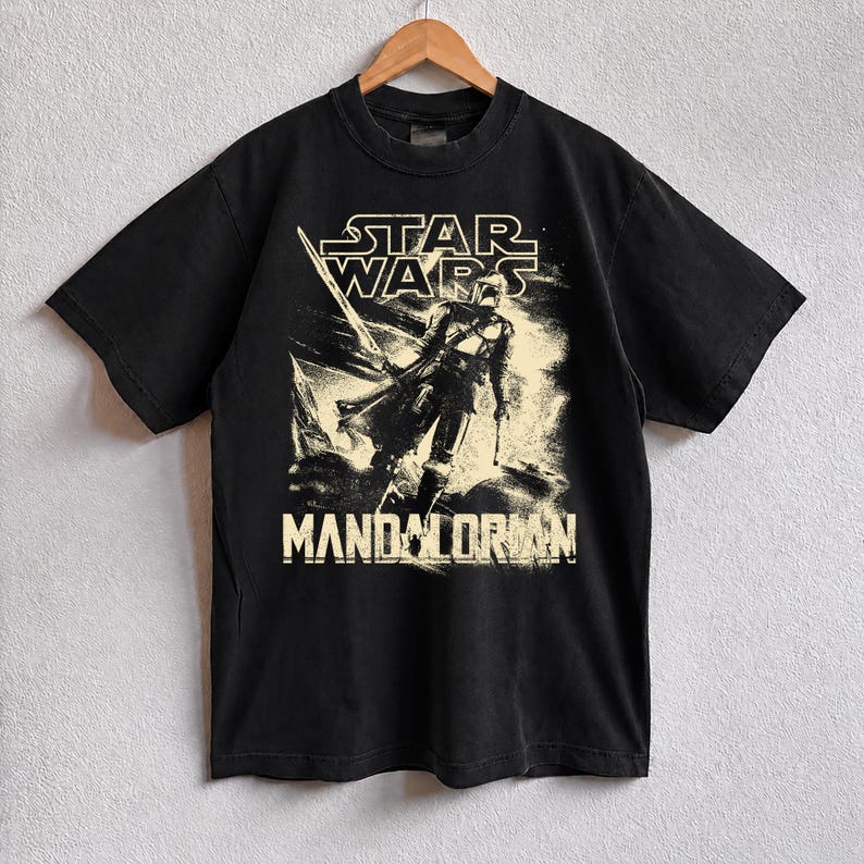 Pu&ograve; includere: T-shirt nera con grafica di The Mandalorian di Star Wars. Il design include il logo di Star Wars e la parola "Mandalorian" in stile vintage usurato. La maglietta &egrave; appesa a una gruccia di legno.