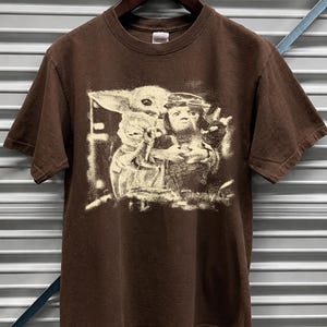 May include: A brown short-sleeved t-shirt with a sepia-toned graphic print of characters from a science fiction franchise. The graphic features a character with large ears and a character with a crown.