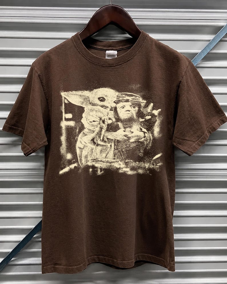 May include: A brown short-sleeved t-shirt with a sepia-toned graphic print of characters from a science fiction franchise. The graphic features a character with large ears and a character with a crown.