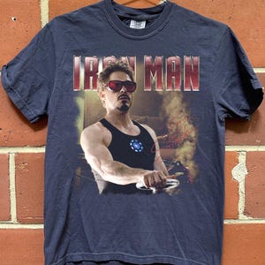 May include: Navy blue t-shirt featuring the text "IRON MAN" in red letters. The shirt displays a graphic of a man wearing sunglasses and a black tank top. The man is holding a silver object. The shirt has a crew neck.