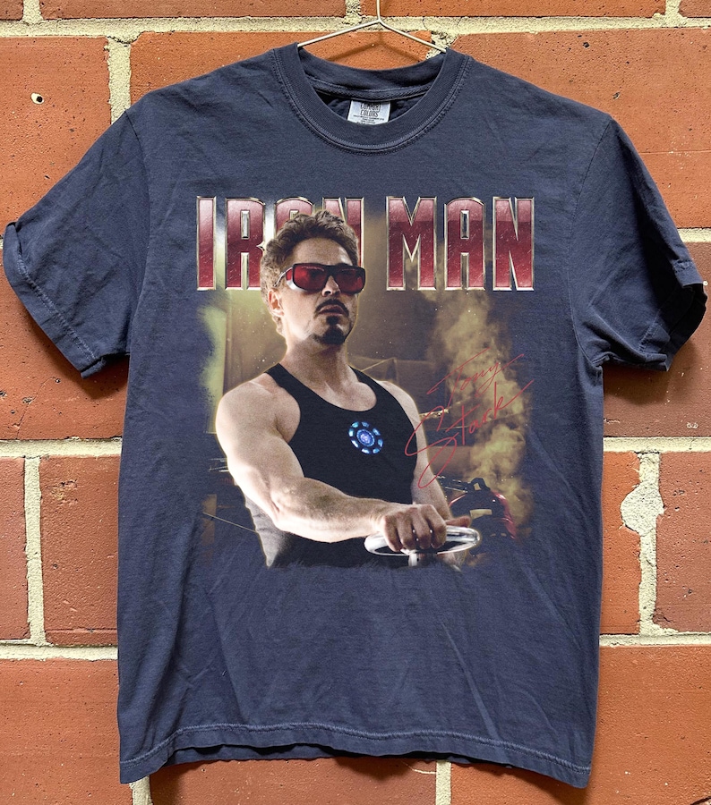 May include: Navy blue t-shirt featuring the text "IRON MAN" in red letters. The shirt displays a graphic of a man wearing sunglasses and a black tank top. The man is holding a silver object. The shirt has a crew neck.
