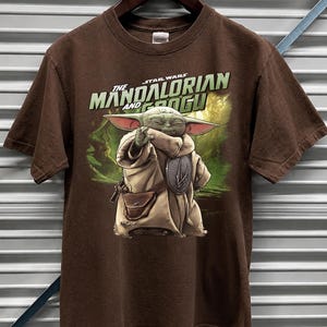 May include: A brown t-shirt with a graphic of Grogu from The Mandalorian. The image shows Grogu in a tan robe with a silver emblem, set against a green and brown background. The text "The Mandalorian and Grogu" is printed above the character.
