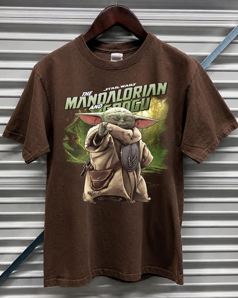 May include: A brown t-shirt with a graphic of Grogu from The Mandalorian. The image shows Grogu in a tan robe with a silver emblem, set against a green and brown background. The text "The Mandalorian and Grogu" is printed above the character.