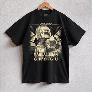 Puede incluir: Camiseta negra con un dise&ntilde;o de Star Wars que presenta a The Mandalorian y Grogu. El dise&ntilde;o incluye el texto "STAR WARS" encima de la imagen y "THE MANDALORIAN AND GROGU" debajo. La camiseta est&aacute; colgada en una percha de madera.