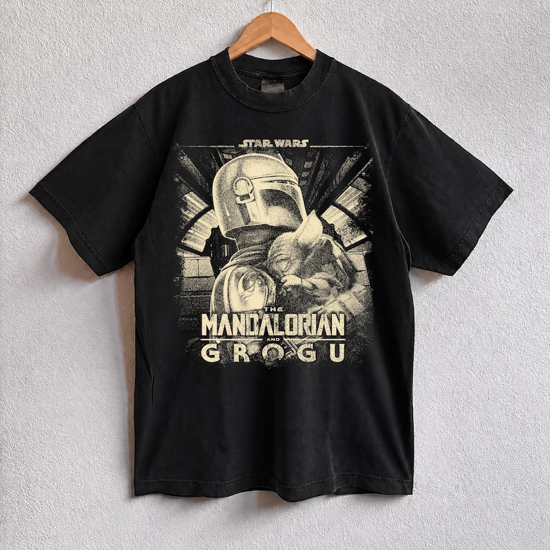 Puede incluir: Camiseta negra con un dise&ntilde;o de Star Wars que presenta a The Mandalorian y Grogu. El dise&ntilde;o incluye el texto "STAR WARS" encima de la imagen y "THE MANDALORIAN AND GROGU" debajo. La camiseta est&aacute; colgada en una percha de madera.