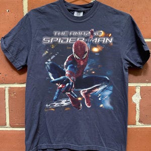 May include: Navy blue t-shirt featuring the Amazing Spider-Man. The graphic shows Spider-Man in a dynamic pose with the text "THE AMAZING SPIDER-MAN" across the top. The shirt is hanging on a metal hanger against a brick wall.