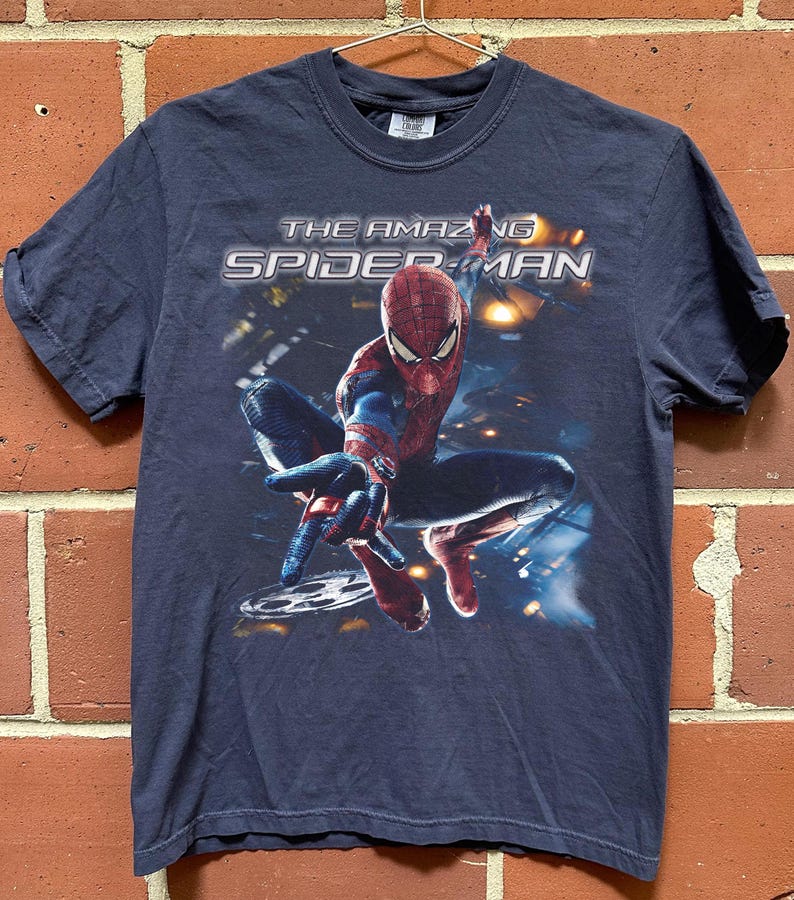 May include: Navy blue t-shirt featuring the Amazing Spider-Man. The graphic shows Spider-Man in a dynamic pose with the text "THE AMAZING SPIDER-MAN" across the top. The shirt is hanging on a metal hanger against a brick wall.