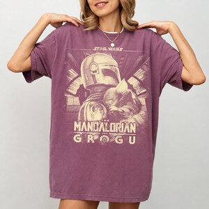 Puede incluir: Una camiseta de color malva con un dise&ntilde;o de Star Wars. El dise&ntilde;o incluye el texto "STAR WARS" sobre una ilustraci&oacute;n de The Mandalorian y Grogu. El gr&aacute;fico es de estilo vintage, con un aspecto desgastado.