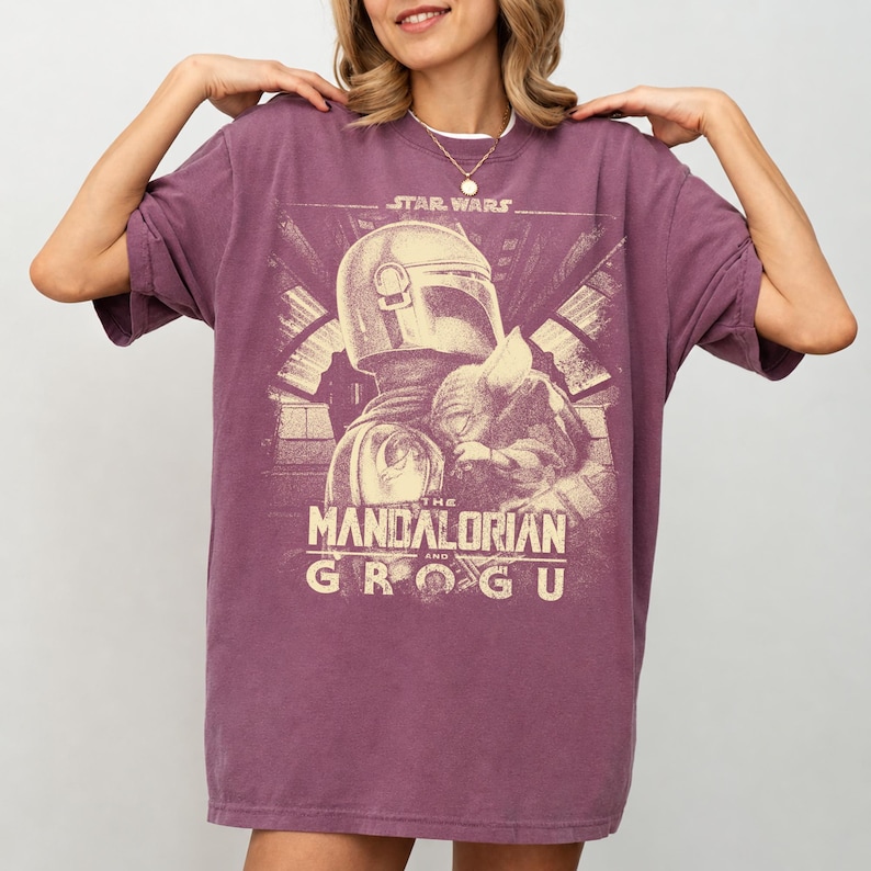 Puede incluir: Una camiseta de color malva con un dise&ntilde;o de Star Wars. El dise&ntilde;o incluye el texto "STAR WARS" sobre una ilustraci&oacute;n de The Mandalorian y Grogu. El gr&aacute;fico es de estilo vintage, con un aspecto desgastado.