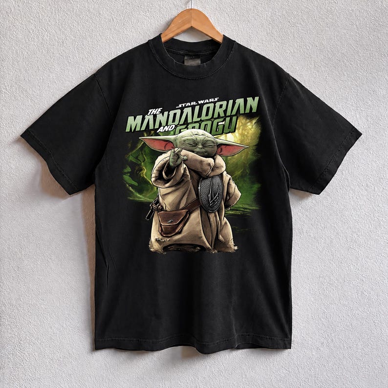 May include: Black t-shirt with a graphic of Grogu from The Mandalorian. The design includes the text "The Mandalorian and Grogu" in green and white. Grogu is shown in a tan robe with a brown satchel and a silver emblem.