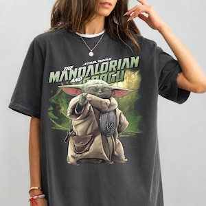 May include: Dark grey t-shirt with a graphic of Grogu from The Mandalorian. The image shows Grogu in a tan robe, holding a silver object, with the text "The Mandalorian and Grogu" in green and white.