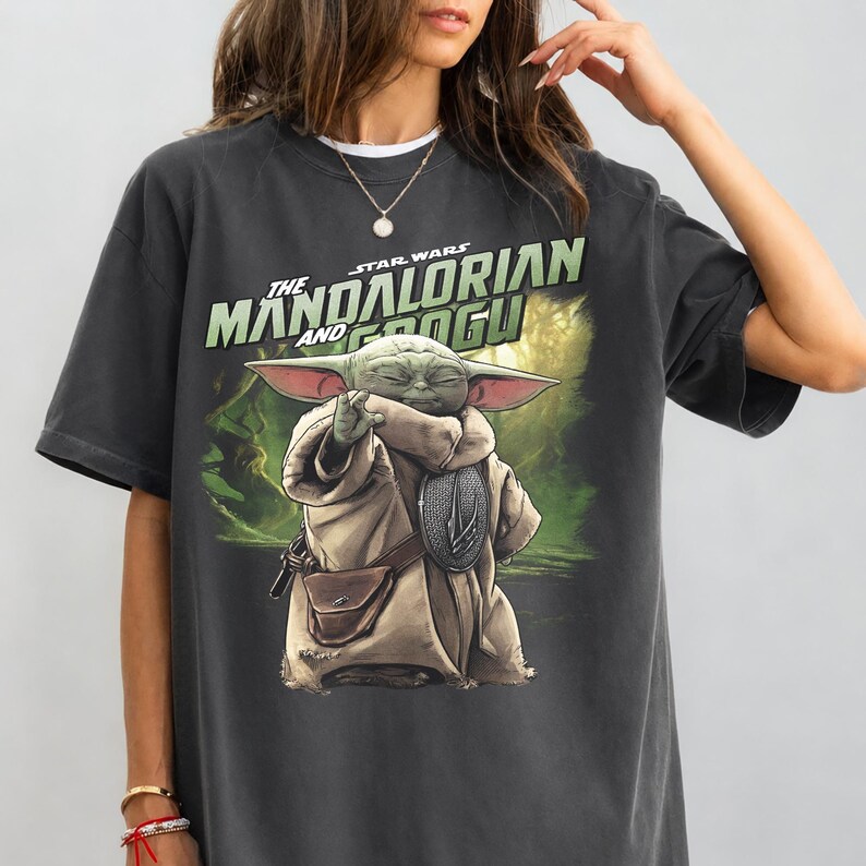 May include: Dark grey t-shirt with a graphic of Grogu from The Mandalorian. The image shows Grogu in a tan robe, holding a silver object, with the text "The Mandalorian and Grogu" in green and white.