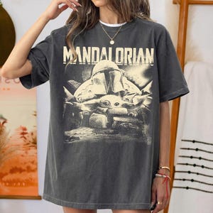 May include: Dark grey t-shirt with the word "MANDALORIAN" and a graphic of the Mandalorian helmet and Grogu. The design is vintage-style. The t-shirt has a relaxed fit.