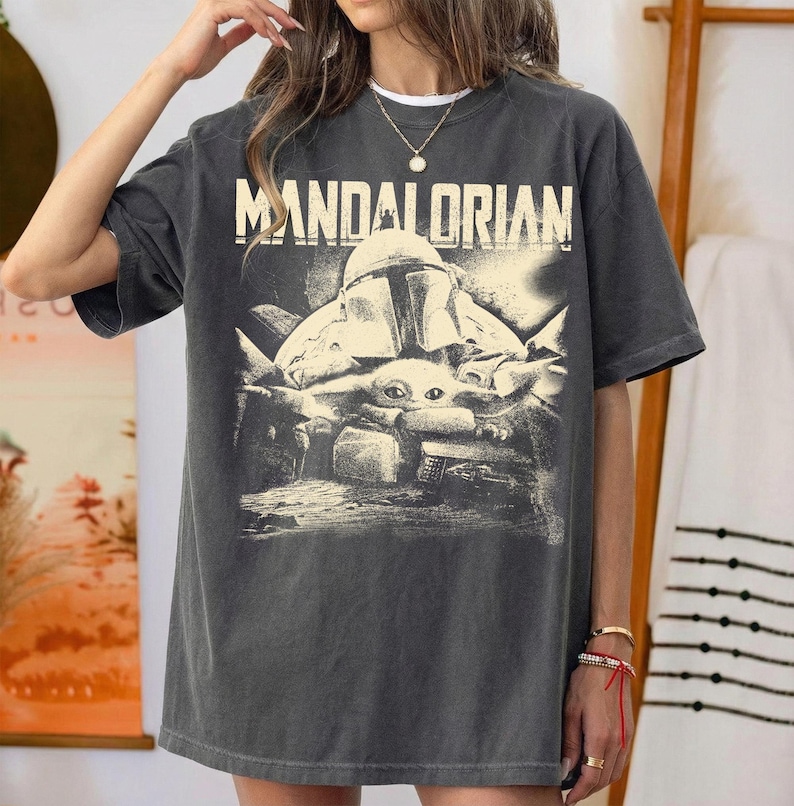 May include: Dark grey t-shirt with the word "MANDALORIAN" and a graphic of the Mandalorian helmet and Grogu. The design is vintage-style. The t-shirt has a relaxed fit.