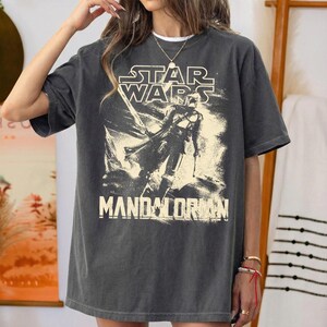 Pu&ograve; includere: T-shirt grigio scuro con il logo di Star Wars e la scritta "Mandalorian" in un font invecchiato. La maglietta presenta una grafica di un personaggio della serie. La maglietta ha una vestibilit&agrave; ampia.