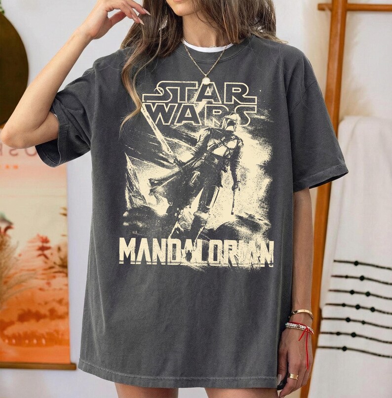 Pu&ograve; includere: T-shirt grigio scuro con il logo di Star Wars e la scritta "Mandalorian" in un font invecchiato. La maglietta presenta una grafica di un personaggio della serie. La maglietta ha una vestibilit&agrave; ampia.