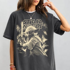 Może przedstawiać: Ciemnoszara koszulka z sepiowym wizerunkiem Yody z Gwiezdnych Wojen, trzymającego miecz świetlny. Logo Star Wars znajduje się nad obrazem. Koszulka ma kr&oacute;tkie rękawy i okrągły dekolt.