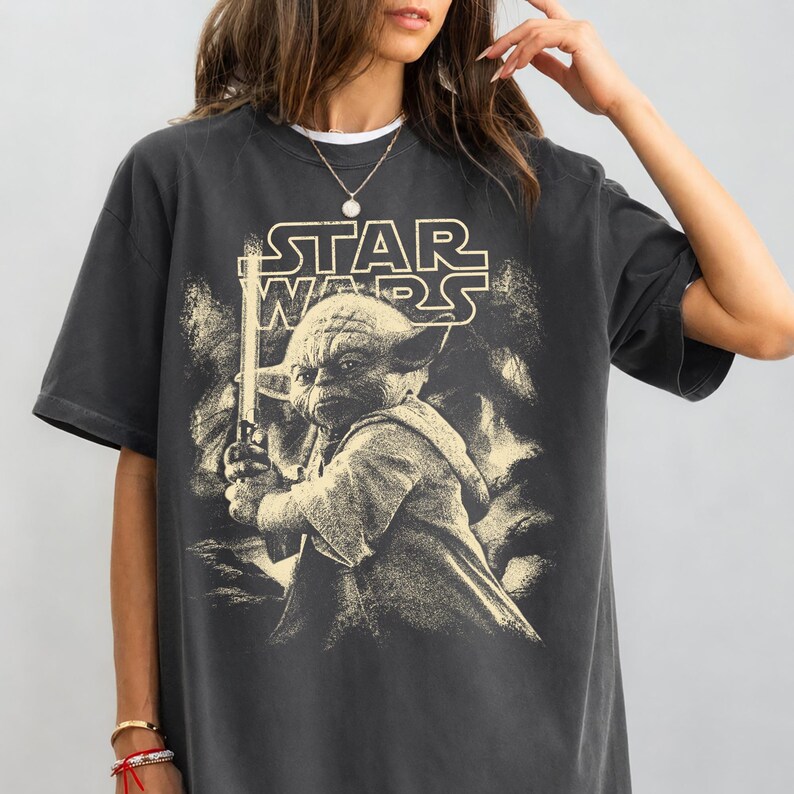 Może przedstawiać: Ciemnoszara koszulka z sepiowym wizerunkiem Yody z Gwiezdnych Wojen, trzymającego miecz świetlny. Logo Star Wars znajduje się nad obrazem. Koszulka ma kr&oacute;tkie rękawy i okrągły dekolt.