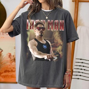 May include: Dark gray t-shirt featuring a graphic of Iron Man, with the text "IRON MAN" in a red and white font. The image shows Tony Stark in sunglasses and a black tank top. The shirt has a vintage, worn look.