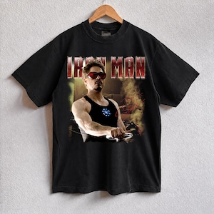 May include: Black t-shirt featuring a graphic of Iron Man, with the text "IRON MAN" in red letters. The image shows a man wearing sunglasses and a black tank top. The shirt is hanging on a wooden hanger.