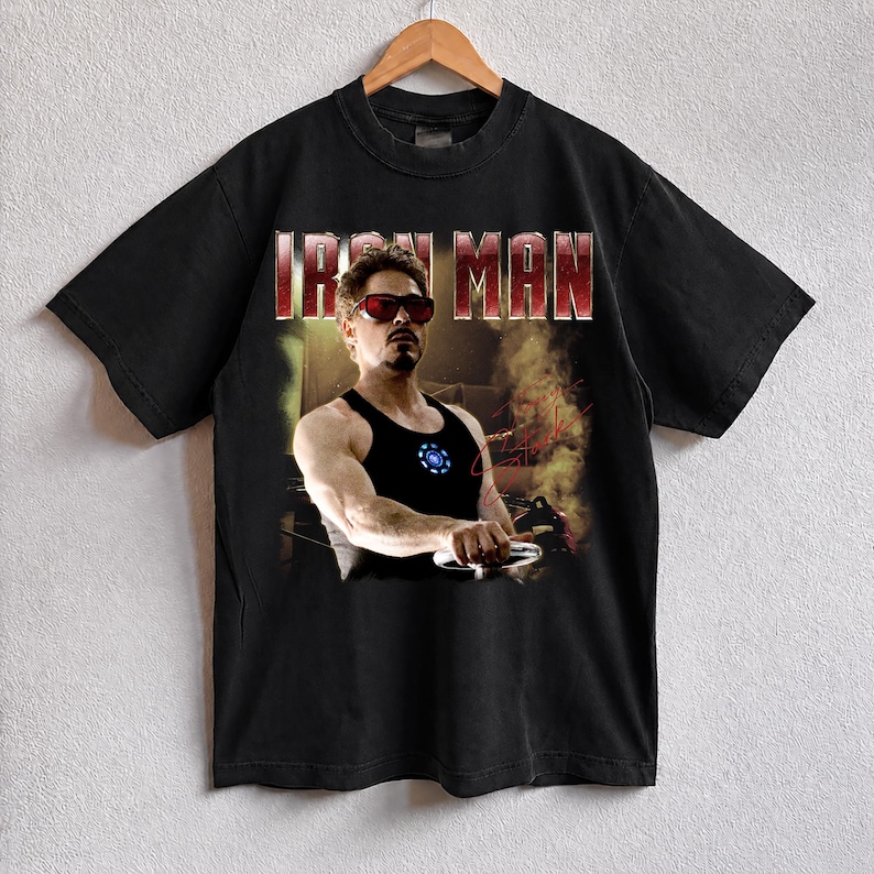 May include: Black t-shirt featuring a graphic of Iron Man, with the text "IRON MAN" in red letters. The image shows a man wearing sunglasses and a black tank top. The shirt is hanging on a wooden hanger.