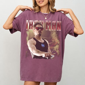 May include: A maroon t-shirt featuring a graphic of Iron Man, with the text "IRON MAN" across the top. The graphic shows a man in sunglasses and a black tank top. The shirt has short sleeves and a crew neck.