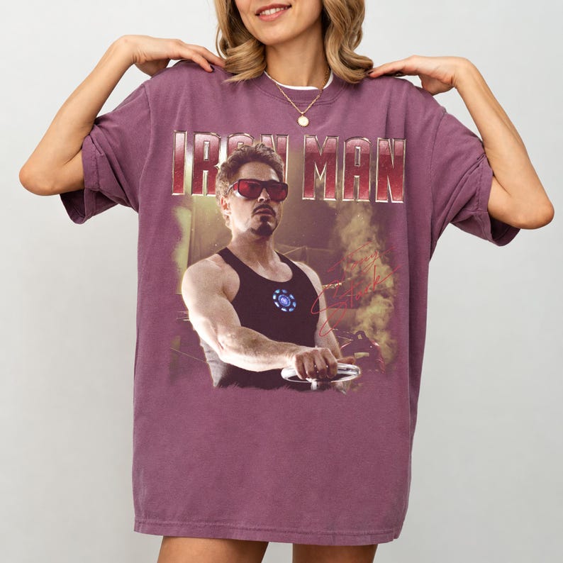 May include: A maroon t-shirt featuring a graphic of Iron Man, with the text "IRON MAN" across the top. The graphic shows a man in sunglasses and a black tank top. The shirt has short sleeves and a crew neck.