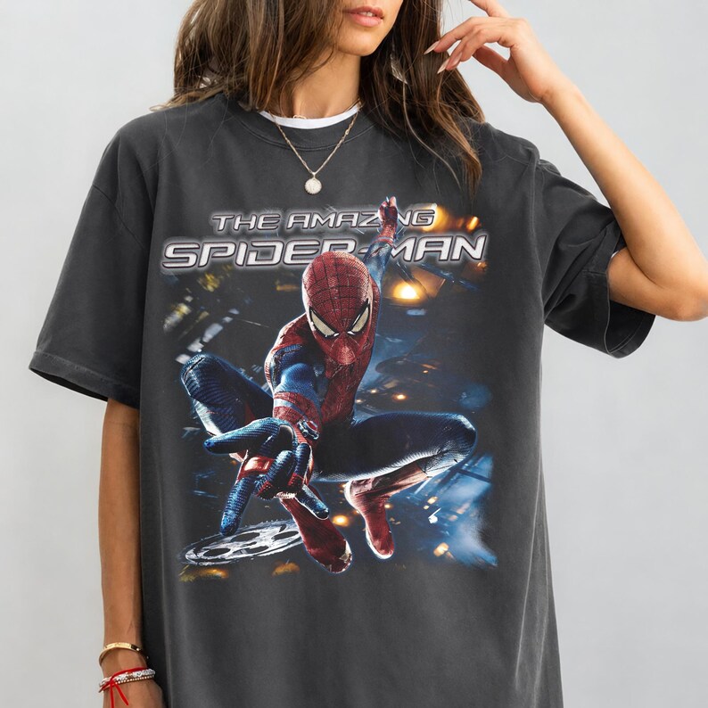 May include: Dark gray t-shirt featuring a graphic of Spider-Man from "The Amazing Spider-Man" movie. The superhero is in a dynamic pose, with the film's title in white and yellow lettering above the image. The shirt has a vintage, worn look.