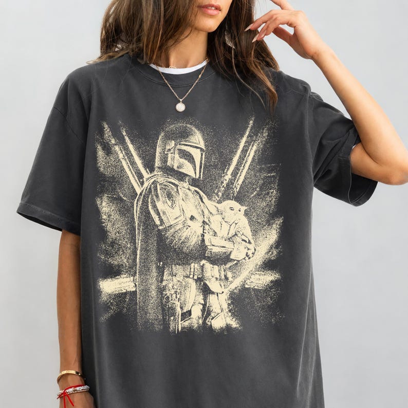 K&ouml;nnte beinhalten: Dunkelgraues T-Shirt mit einem verblichenen Aufdruck eines Mandalorianers, der eine kleine Kreatur h&auml;lt. Das Design zeigt eine Figur mit Helm und Umhang sowie zwei vertikale, lichtschwertartige Elemente. Vintage-Stil.