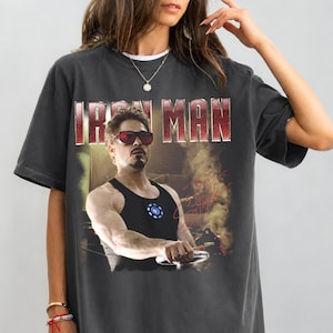May include: Dark gray t-shirt featuring a graphic of Iron Man, with the text "IRON MAN" in a red and white font. The image shows a man wearing sunglasses and a black tank top. A signature is visible on the right side of the graphic.
