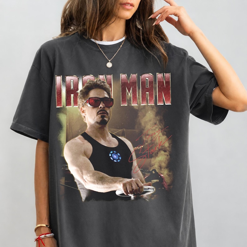 May include: Dark gray t-shirt featuring a graphic of Iron Man, with the text "IRON MAN" in a red and white font. The image shows a man wearing sunglasses and a black tank top. A signature is visible on the right side of the graphic.