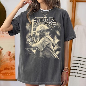 Może przedstawiać: Ciemnoszara koszulka z grafiką Star Wars. Projekt zawiera logo Star Wars i wizerunek Yody trzymającego miecz świetlny. Koszulka ma wygląd vintage.