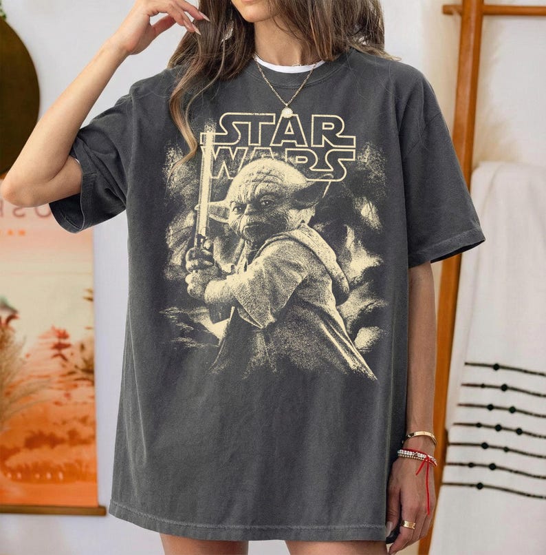 Może przedstawiać: Ciemnoszara koszulka z grafiką Star Wars. Projekt zawiera logo Star Wars i wizerunek Yody trzymającego miecz świetlny. Koszulka ma wygląd vintage.