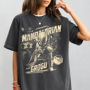 Pu&ograve; includere: T-shirt grigio scuro con grafica della serie Star Wars, "The Mandalorian and Grogu". Il design include il personaggio di Mandalorian e Grogu, con testo in stile vintage. La maglietta ha maniche corte e girocollo.