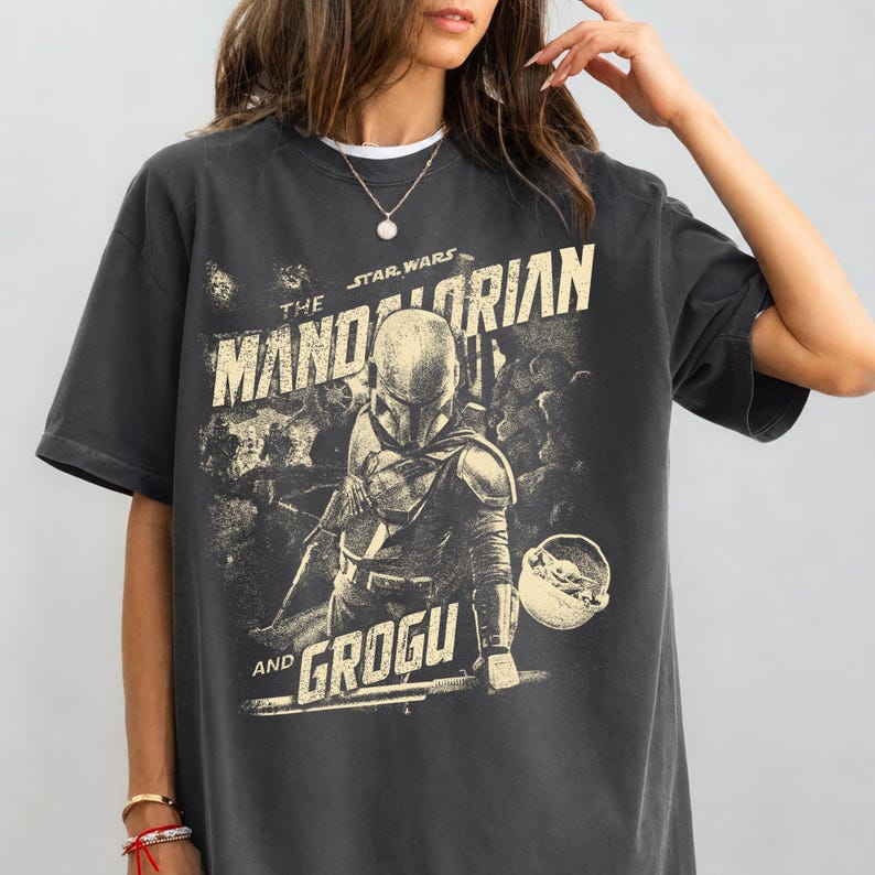 Pu&ograve; includere: T-shirt grigio scuro con grafica della serie Star Wars, "The Mandalorian and Grogu". Il design include il personaggio di Mandalorian e Grogu, con testo in stile vintage. La maglietta ha maniche corte e girocollo.