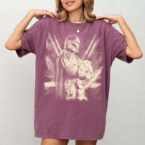 K&ouml;nnte beinhalten: Ein malvenfarbenes T-Shirt mit einem verblichenen Aufdruck eines Charakters mit Helm, der eine kleine Kreatur h&auml;lt. Das Design ist in einem hellen Beige gehalten, im Vintage-Stil. Das Shirt hat kurze &Auml;rmel und eine lockere Passform.
