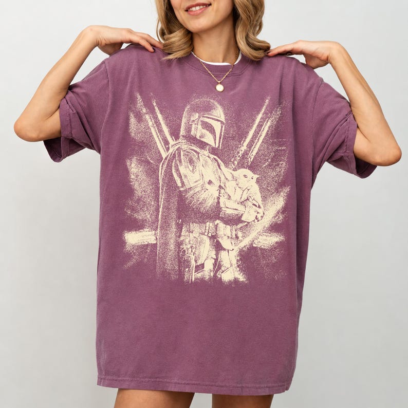 K&ouml;nnte beinhalten: Ein malvenfarbenes T-Shirt mit einem verblichenen Aufdruck eines Charakters mit Helm, der eine kleine Kreatur h&auml;lt. Das Design ist in einem hellen Beige gehalten, im Vintage-Stil. Das Shirt hat kurze &Auml;rmel und eine lockere Passform.
