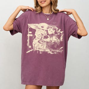 May include: A plum-coloured, short-sleeved oversized t-shirt with a faded graphic print. The print shows a character with large ears and a figure wearing goggles. The shirt has a crew neck and a relaxed fit.