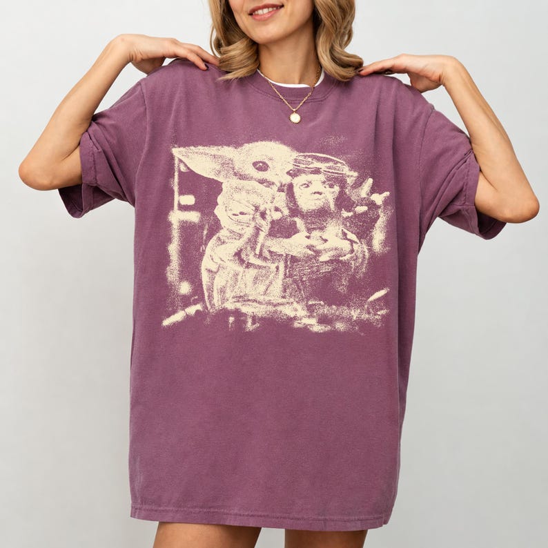 May include: A plum-coloured, short-sleeved oversized t-shirt with a faded graphic print. The print shows a character with large ears and a figure wearing goggles. The shirt has a crew neck and a relaxed fit.